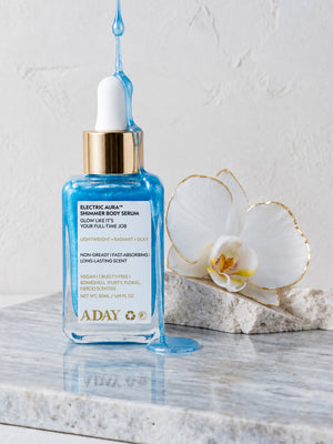ADAY Electric Aura Shimmer Body Serum, a 50 ml blue glass dropper bottle on marble beside a white orchid